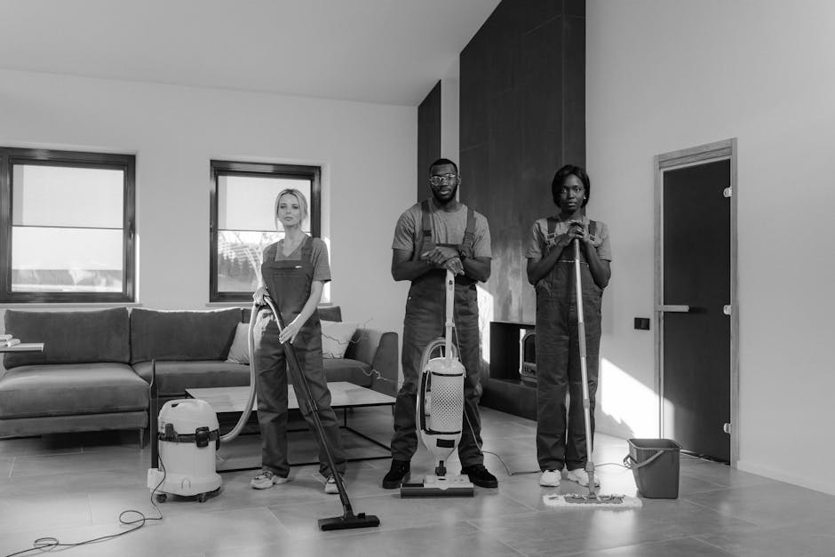 Three professional cleaners from Hither Green Carpet Cleaners are positioned in a modern living room, actively engaged in surface cleaning tasks. The room features a polished tiled floor, a contempora