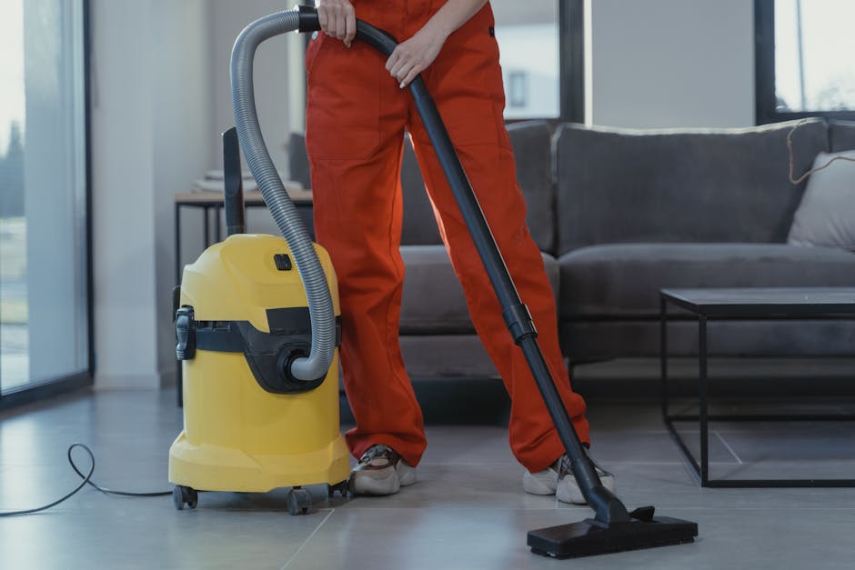 A person dressed in orange overalls vacuuming a light-colored tiled floor in a modern living room. The vacuum cleaner, yellow with a black handle and hose, is positioned in front of the individual, wh